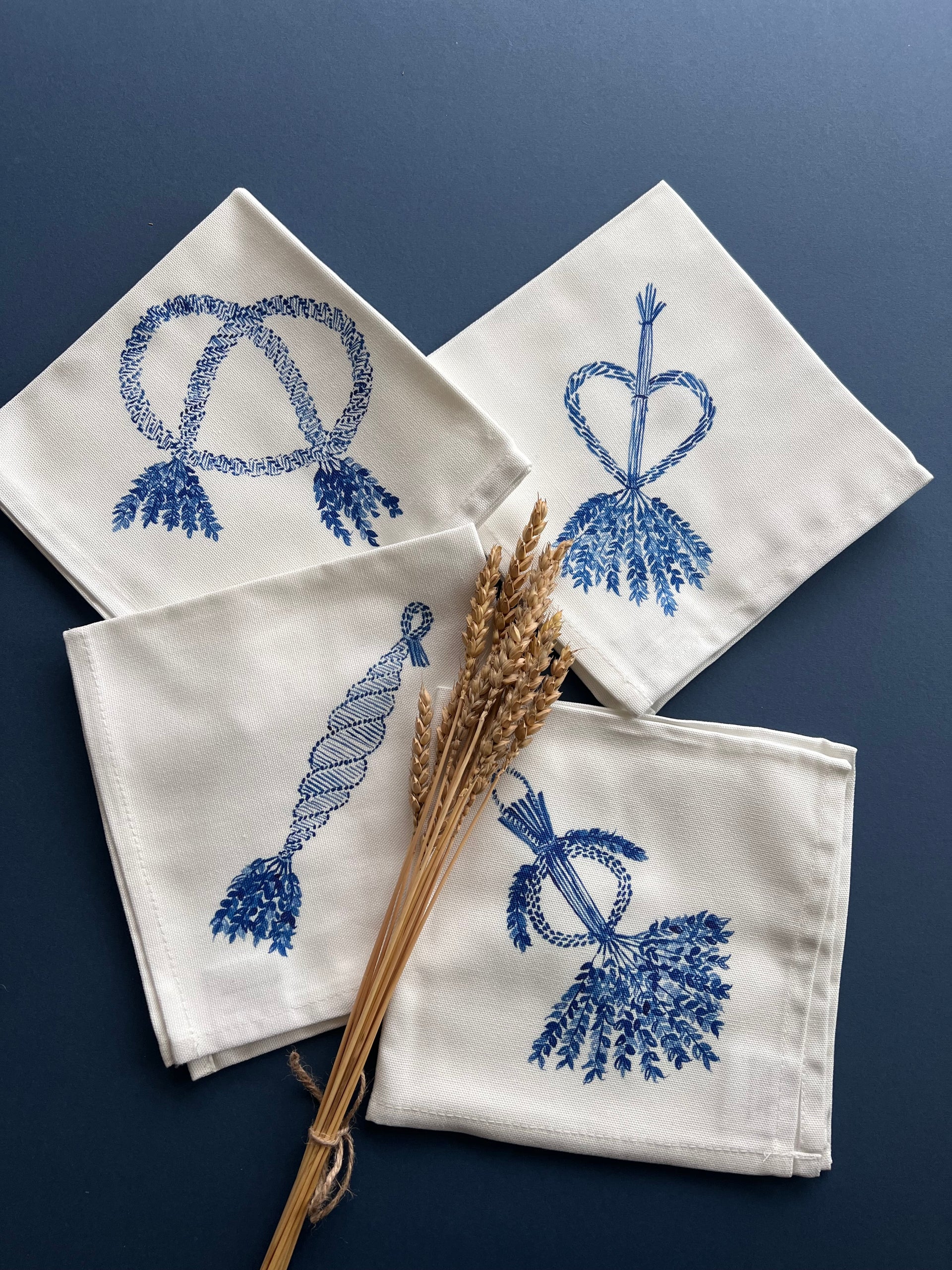 Set of four square white napkins printed with corn dolly images in blue, each napkins shows a different design of corn dolly.